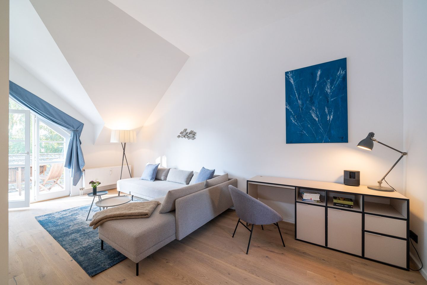 Auf dem Bild ist der Wohnraum des Apartments Blausand zu sehen. Auf der rechten Seite steht eine beige Couch in L-Form, mit blauen Kissen darauf. Vom Wohnzimmer aus tritt man auf den kleinen Balkon. Zudem ist das Wohnzimmer mit einem kleinen Arbeitstisch ausgestattet.
