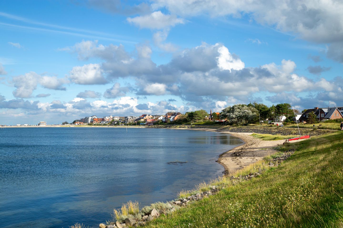 Küstenpanorama von Wittdün auf Amrum mit Blick über das ruhige Wasser auf die Skyline des Inselortes, gesäumt von einem schmalen Sandstrand und grünen Deichwiesen unter blauem Wolkenhimmel.