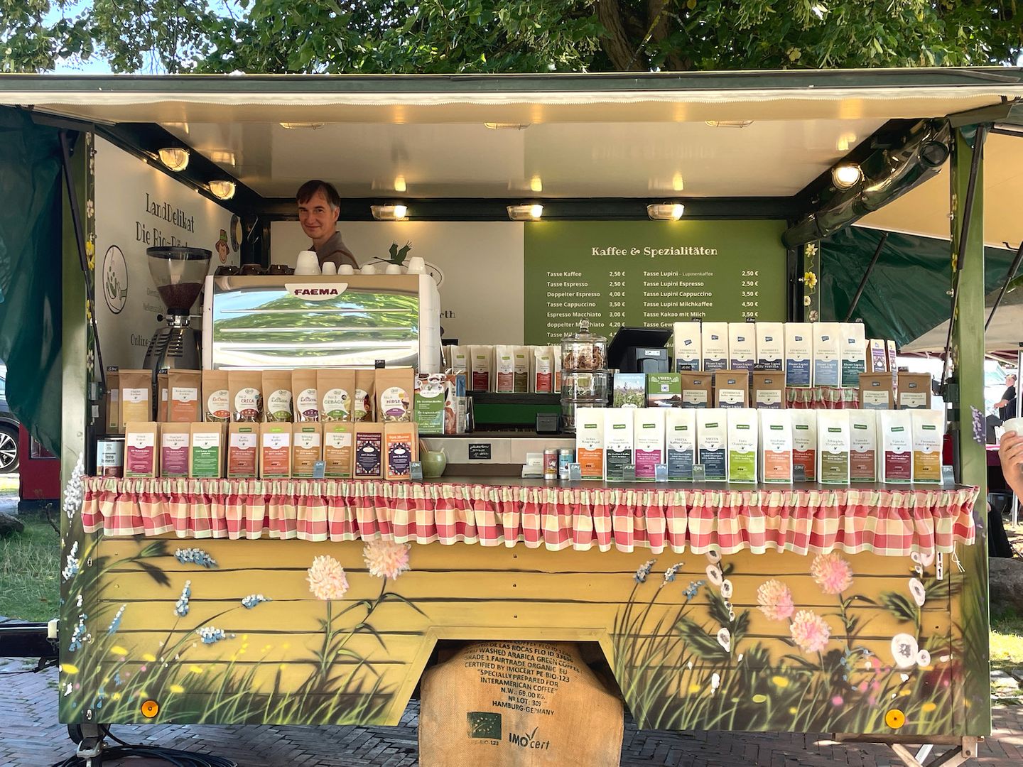 Kaffeestand auf einem Markt, Blick in den Stand mit Mann an der Kaffeemaschine, verschiedene Kaffeetüten im Vordergrund